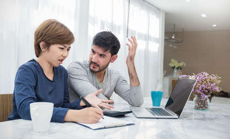 Worried couple paying bills online via laptop in living room at home. Young man and woman at desk...
