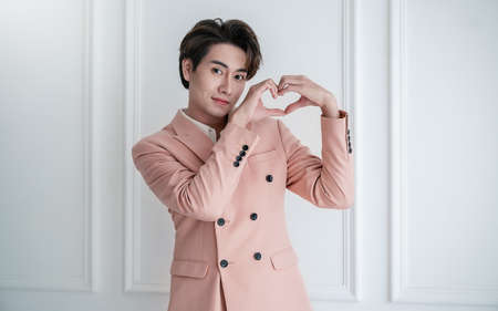 Portrait of young handsome smile asian business man in pink suit hands gesture in heart shape. Smile face asian guy model. Japanese or korean boy lifestyle, happy man valentines day, love conceptの写真素材