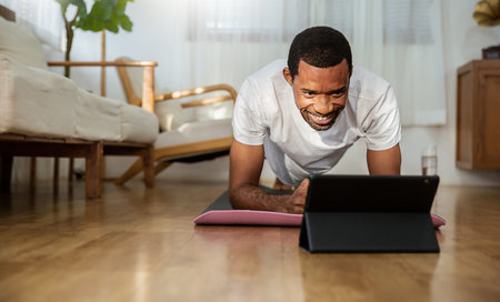 Portrait of young African American black man warm up exercise workout yoga plank position at home living room. Yoga for beginner. Guy watching online tutorials on tablet, training, healthy stay homeの写真素材