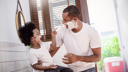 Portrait of happy  African American black father son apply shaving foam on face while looking in reflection mirror morning routine. Man shaving with razor, hygiene body care, family lifestyle conceptの写真素材