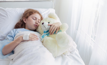 Little caucasian girl not well recover lying in hospital bed with teddy bear. Student girl reading book while admission.Health care and medical education concept.の写真素材