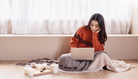 Asian nerd glasses woman work with virtual computer on the floor. Warm woolen sweater in cold autumn or winter weekend, warm drink. Sme small business work from home online tecの写真素材