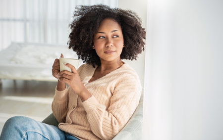 Portrait of young beautiful African American black woman hands holding coffee cup morning spring time in white bedroom. Happy cheerful relaxing in winter. Wakeup university lifestyle conceptの写真素材