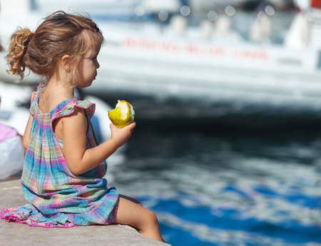 Little blond girl sits on the dock with pear in her hand.の写真素材