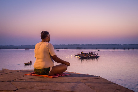 VARANASI, INDIA - 23 FEBRUARY 2015: Indian man meditates next to sacred Ganges river on  Varanasi ghat while people perform puja on boats in background.のeditorial素材