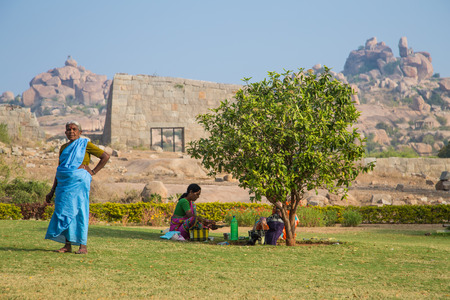 HAMPI, INDIA - 30 JANUARY 2015: Elderly woman stands on small grass field next to tree and family.のeditorial素材