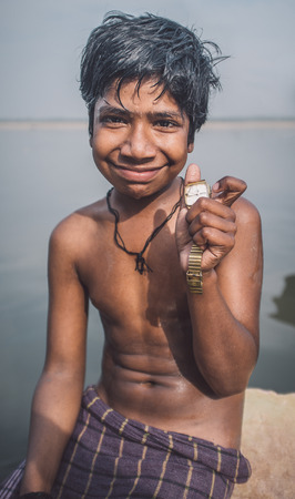 VARANASI, INDIA - 25 FEBRUARY 2015: Indian boy sits shirtless on rock in Ganges river and shows watch he found. Post-processed with grain, texture and colour effect.のeditorial素材