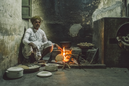 GODWAR REGION, INDIA - 12 FEBRUARY 2015: Indian man dressed in traditional clothes makes chapati on open fire in old kitchen. Post-processed with grain, texture and colour effect.のeditorial素材