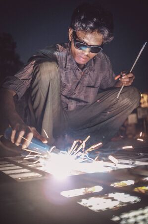 KAMALAPURAM, INDIA - 02 FABRUARY 2015: Indian worker welds heavy metal parts on street in dusk. Post-processed with grain, texture and colour effect.の写真素材