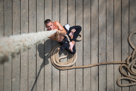 Young shirtless athlete man sitting on the plank floor holding the rope as part of crossfit...