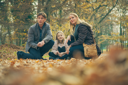 Cheerful family of three kneel on park ground covered with leaves on an autumn day.の写真素材