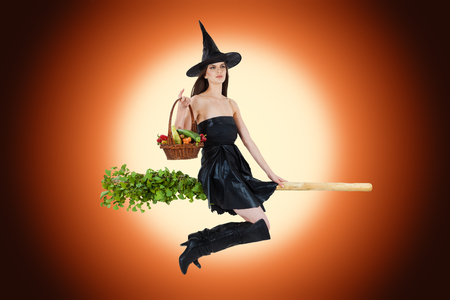 Young woman dressed as a witch carrying vegetable basket and riding a broom. Radial orange background.の写真素材