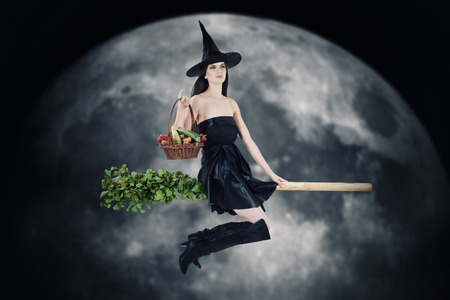 Young woman dressed as a witch carrying vegetable basket and riding a broom. Blurred moon in background.の写真素材