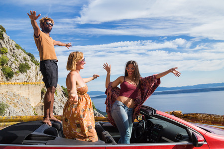 Group of happy young people standing in the red convertible car and waving.の写真素材