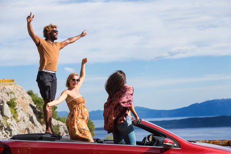 Group of happy young people standing in the red convertible car and waving.の写真素材