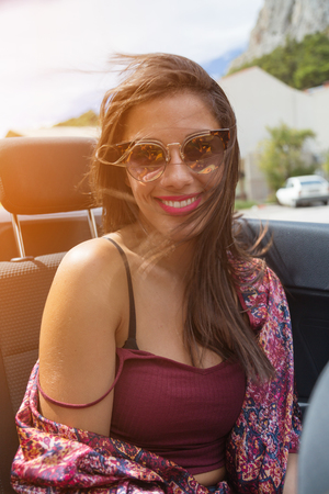 Happy brunette girl sitting and smiling in the back seat of convertible car. Sun effect applied.の写真素材