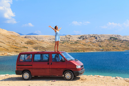 Beautiful tanned girl in a blue dress standing on a rooftop of red van and spreading arms.の写真素材