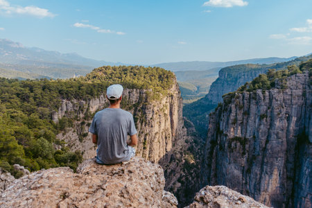 A guy in a cap and T-shirt sits looking at the panoramic landscape of lazi Kanyonu (Eagles Canyon) and Bilgelik Vadisi (Wisdom Valley). Located in Koprulu Canyon National Park, Antの写真素材