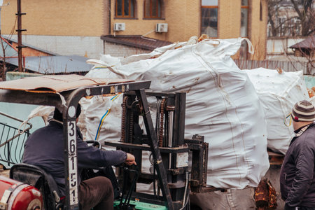 A forklift unloads large bags of firewood from a truck and stacks them along the fence on a cloudy winter's dayの写真素材