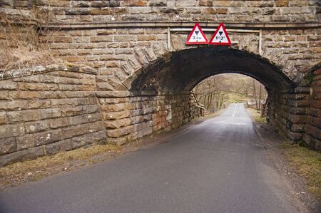Old stone railway bridge with low warning signs over a country laneの写真素材