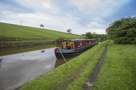 Narrowboat moored up in English rural countryside scenery on British waterway canal during overcast cloudy dayの写真素材