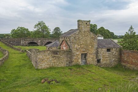 Old abandoned derelict mill stone building in rural countryside setting with overgrown bridgeの写真素材