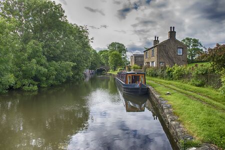 Narrowboat moored up in English rural countryside scenery on British waterway canal during overcast cloudy dayの写真素材
