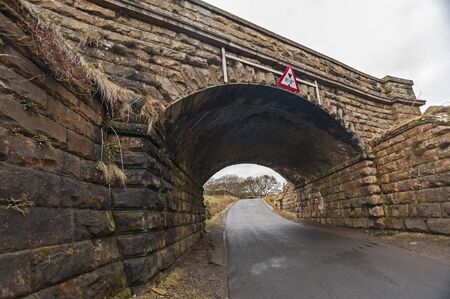 Old stone railway bridge with low warning signs over a country lane
