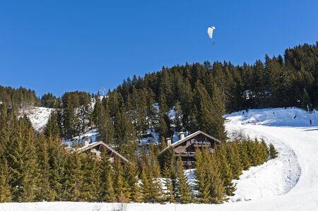 Paraglider flying over a snow covered alpine mountain resort with chalets and trees