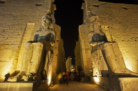 Large statue and hieroglypic carvings of Ramses II at the entrance to ancient egyptian Luxor temple lit up in nightの写真素材