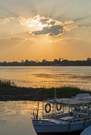 Wooden boats moored on tropical river bank at dusk sunset with sun and cloud in orange skyの写真素材