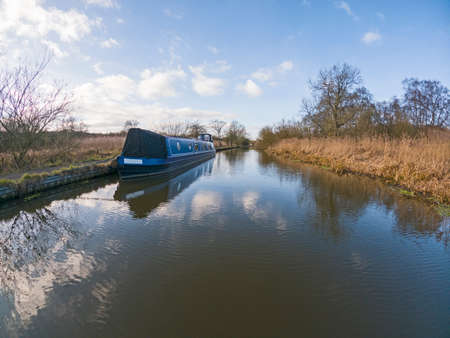 Narrowboat moored up in English rural countryside scenery on British waterway canal with cloudy blue skyの写真素材