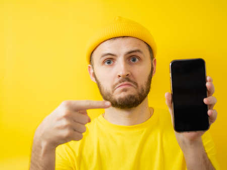 Portrait of an excited young man, pointing at a mobile phone with his index finger. Isolated on a yellow backgroundの写真素材