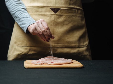 Woman hand sprinkle raw chicken fillet by condiment.
