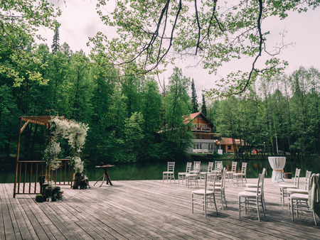 Romantic rustic wedding ceremony near river. White wooden chairs and flower arch.の写真素材