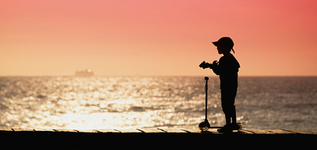 Silhouette child with scooters and binoculars at sunset by the oceanの写真素材