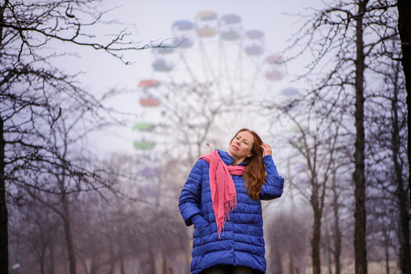 girl in a red scarf stands in the park on an autumn colorful afternoon opposite the ferris wheel and enjoys natureの写真素材