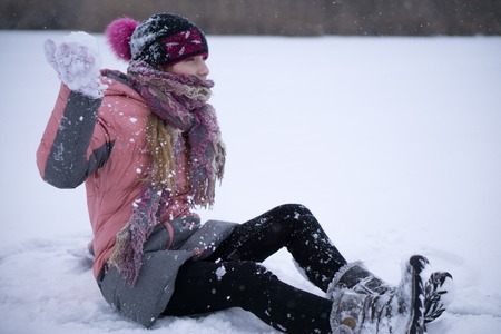 closeup shot of a young teenage girl sit alone on a frozen ground lake in snow and ready to throw snowball in her hand face head wrapped in scarf.の写真素材