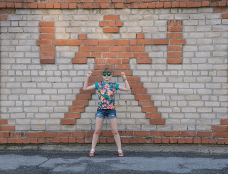 one young teenage girl in mirrored glasses goggles standing next to the brick wall hands up pretending lifting barbell doing workout wearing flowered t-shirt and jeans shorts copying figure of athlete bodybuilder powerlifter with barbell made of bricks onの写真素材