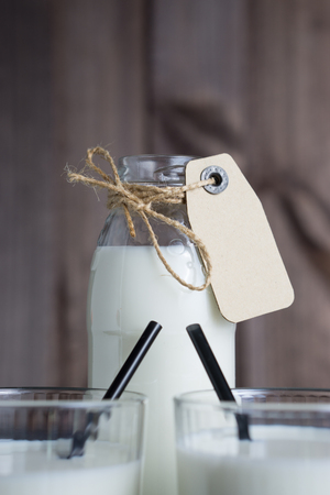 closeup shot bottle of milk with carton label tag with two transparent glasses, black straw on dark wooden backgroundの写真素材