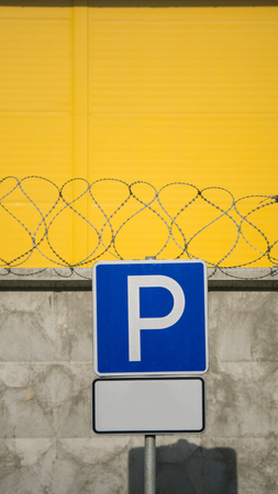 Parking Sign - Blue road sign with letter P on rectangular plate against a concrete fence and yellow wall with barbed wire. empty space for time or other conditionsの写真素材