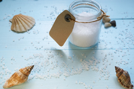 small jar of sea salt with carton cardboard tag on blue desk with a few seashells and scattered salt all over the desk. low angle view with lot of copy spaceの写真素材