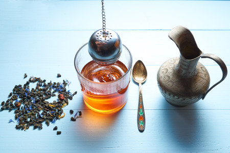 glass of tea with jug, teaspoon and tea leaves on bright blue desk. visible waves circles on tea surface. strainer out of glass for better brew. high angle shotの写真素材
