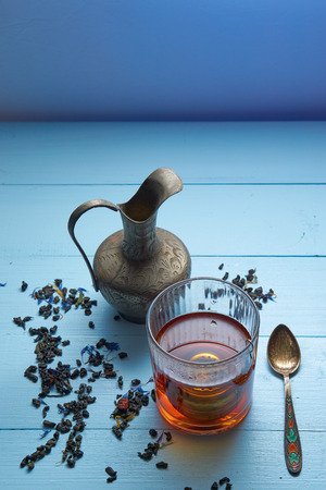 vertical shot of herbal tea glass with strainer, middle east decorative jug, vivid teaspoon and tea leaves on blue background. high angle view. lot of copy space for caption headerの写真素材