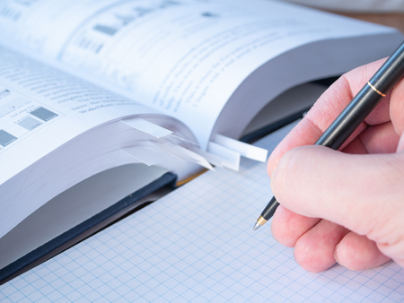 side view of a student hand with pen above empty sheets of squared notepad and blurred book with bookmarks on background. close-up low angle shotの写真素材