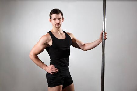 Young strong pole dancer smiling man in black clothes on pylon. Neutral background.