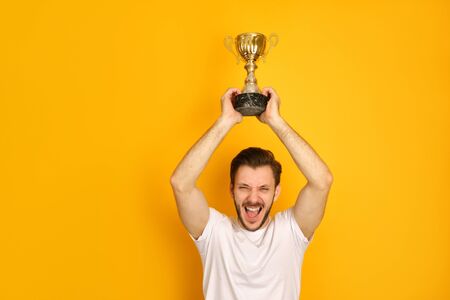 A young sportsman on yellow background, smiling happily, shouting out loud, holding a golden prize over his head, glad to win, wearing a white T-shirtの写真素材