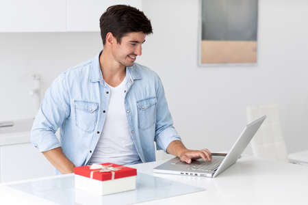 Young handsome guy is choosing a gift for his girlfriend online. Man with laptop at the table at home.の写真素材