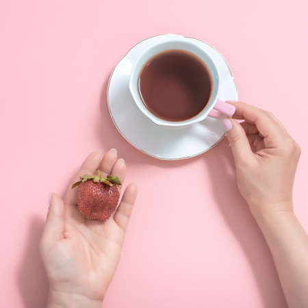 women's hands with a Cup of tea and strawberries on a pink background. white Cup of tea on a white saucer. top viewの写真素材