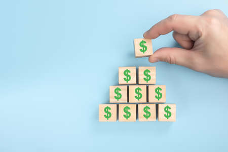 Hand putting wooden cube block on top pyramid of dollars. Wood block cubic stacking up with green...